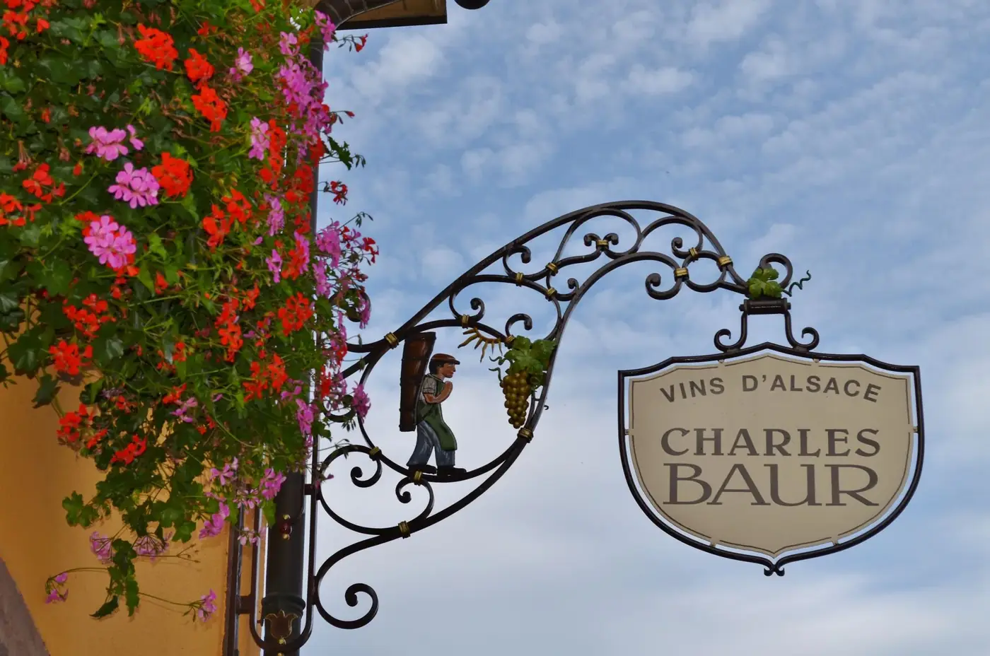 Ornate wine shop sign in Alsace, France with vibrant flowers and clear sky.