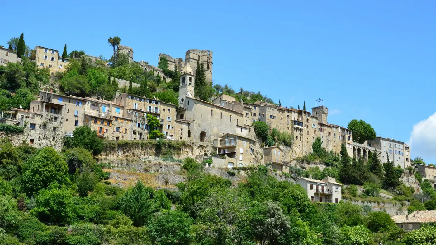 Explore the historic stone architecture of Montbrun-les-Bains with lush greenery under a blue sky.