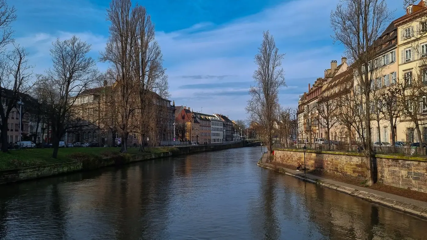 Serene view of Strasbourg canal flanked by historic buildings and bare trees under a bright winter sky.