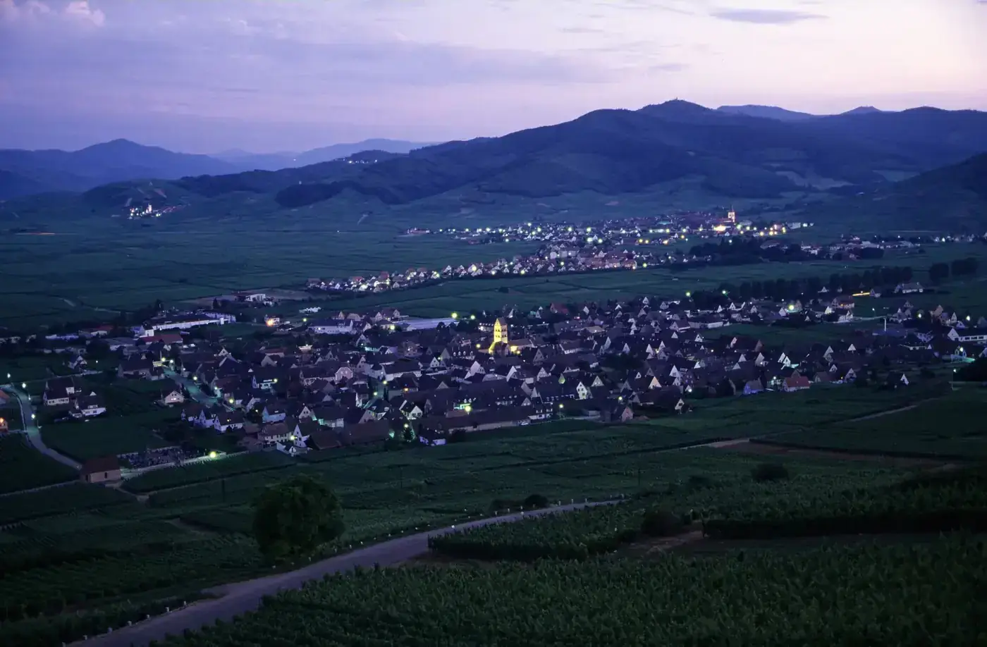 A charming hillside village in Alsace surrounded by vineyards at twilight, with mountain views.
