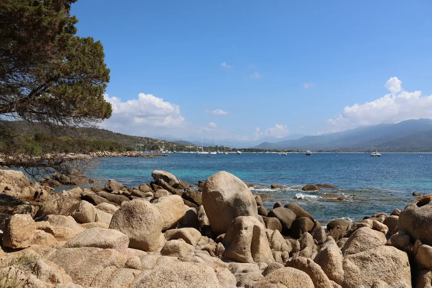 Scenic seascape of rocky shore, clear blue water, and sailboats.