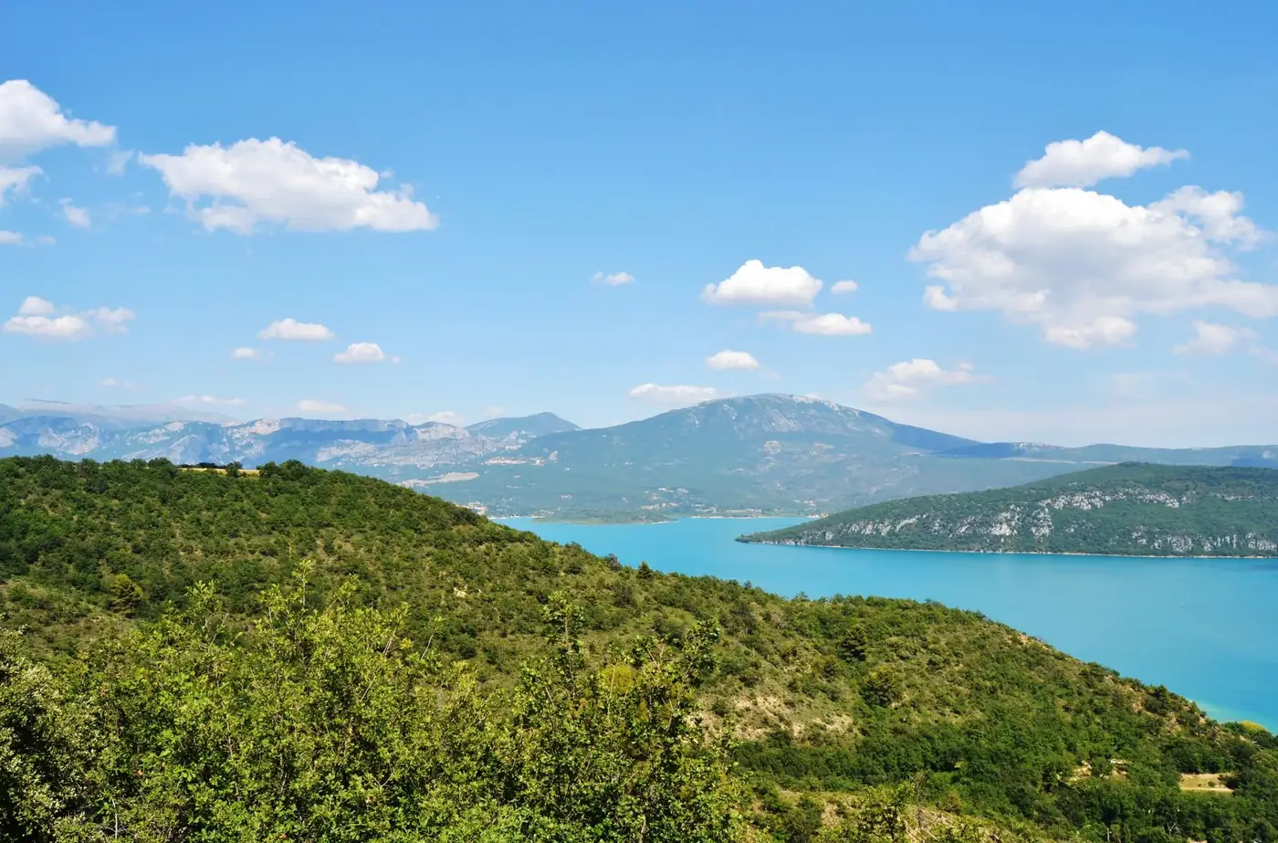 A breathtaking view of Lake Sainte-Croix surrounded by lush hills and distant mountains under a bright blue sky.